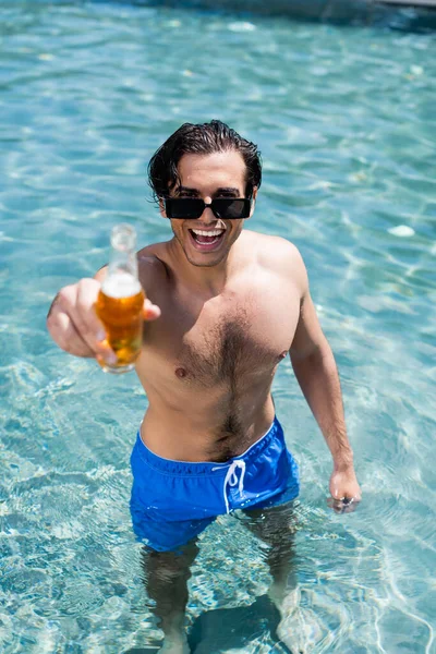 Uomo eccitato in costume da bagno guardando la fotocamera mentre tiene in mano una bottiglia di birra in piscina — Foto stock