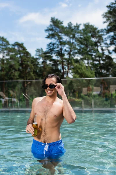 Uomo sorridente regolazione occhiali da sole mentre in piedi con birra in piscina — Foto stock