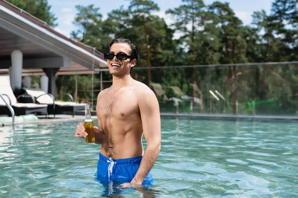 Homme torse nu heureux dans des lunettes de soleil détente dans la piscine avec bouteille de bière — Photo de stock