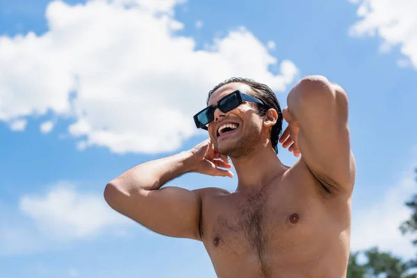 Vista basso angolo di felice uomo senza camicia in occhiali da sole contro cielo nuvoloso blu — Foto stock
