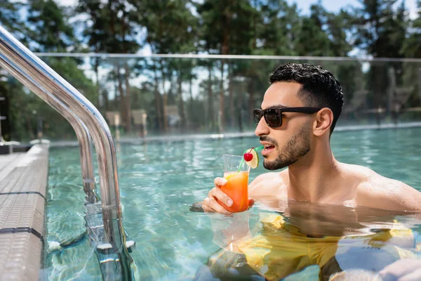 Joven musulmán hombre en gafas de sol beber cóctel en la piscina - foto de stock