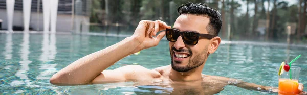 Gioioso uomo arabo che regola gli occhiali da sole mentre si rilassa in piscina con cocktail, banner — Foto stock