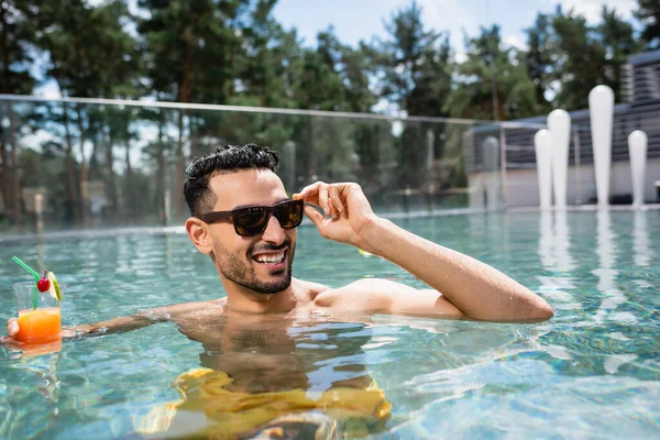 Homem árabe satisfeito tocando óculos de sol enquanto descansa com coquetel na piscina — Fotografia de Stock