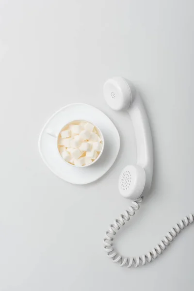 Вид зверху солодких цукрових кубиків у чашці з тарілкою біля ретро телефону на білому — стокове фото