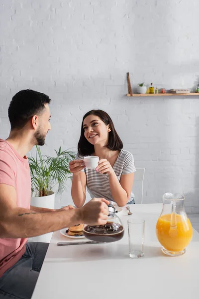 Улыбающаяся молодая женщина с чашкой кофе смотрит на парня с кофейником на кухне — стоковое фото