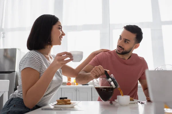 Sonriente joven con taza tocando novio con cafetera en cocina - foto de stock