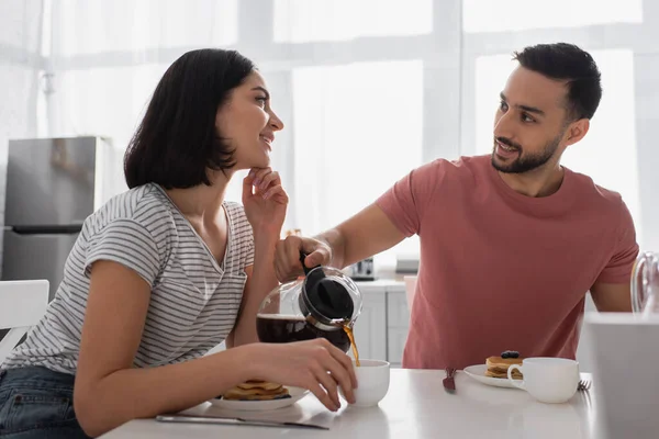 Улыбающаяся молодая женщина с рукой возле лица, глядя на бойфренда, наливающего кофе в чашку на кухне — стоковое фото