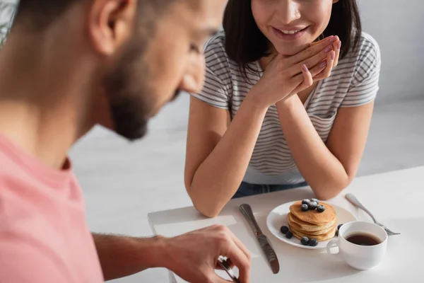 Mujer joven con las manos cerca de sentarse cerca de la cara borrosa novio comiendo panqueques en la cocina — Stock Photo