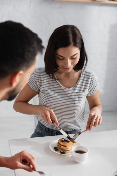 Young woman eating pancakes near blurred boyfriend in kitchen — Stock Photo