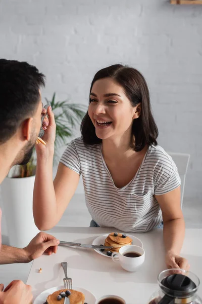 Positive jeune femme nourrir petit ami avec des morceaux de crêpes sur la fourchette dans la cuisine — Photo de stock