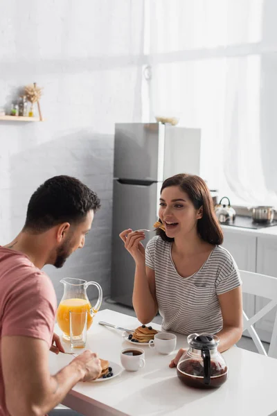 Jovem feliz comendo pedaços de panquecas em garfo com namorado na cozinha — Fotografia de Stock