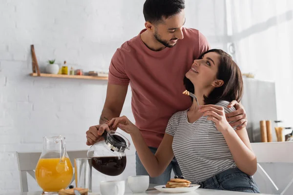 Happy young man hugging girlfriend with pieces of pancakes on fork and pouring coffee from pot to cup in kitchen — Stock Photo