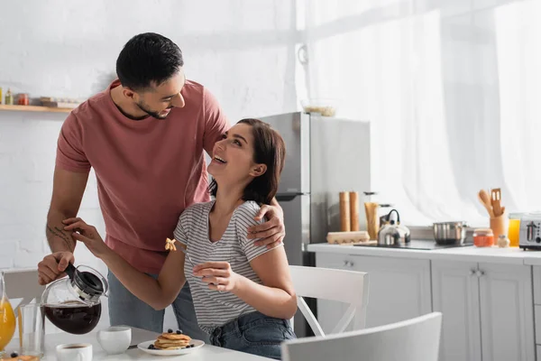 Smiling young man hugging girlfriend with pieces of pancakes on fork and pouring coffee from pot to cup in kitchen — Stock Photo