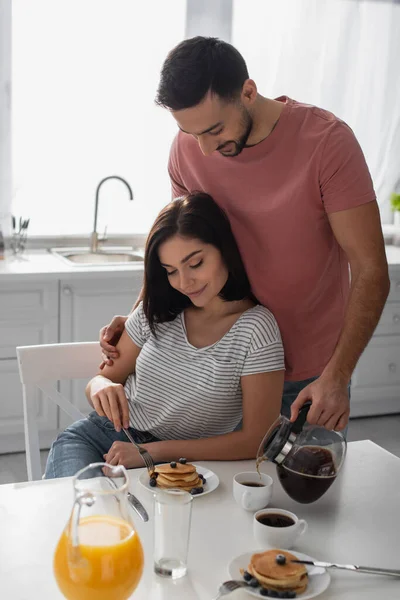 Sonriente joven abrazando a su novia comiendo panqueques y vertiendo café de olla en taza en la cocina - foto de stock