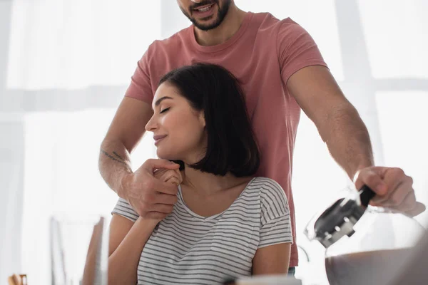 Young man hugging girlfriend with closed eyes and pouring coffee from pot in kitchen — Stock Photo