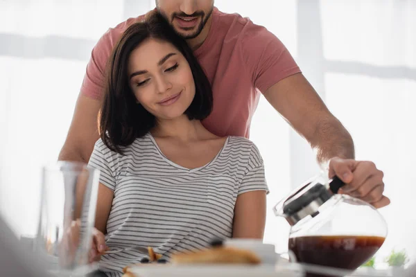 Joven abrazando a su novia con el desayuno y vertiendo café de olla en taza en la cocina - foto de stock
