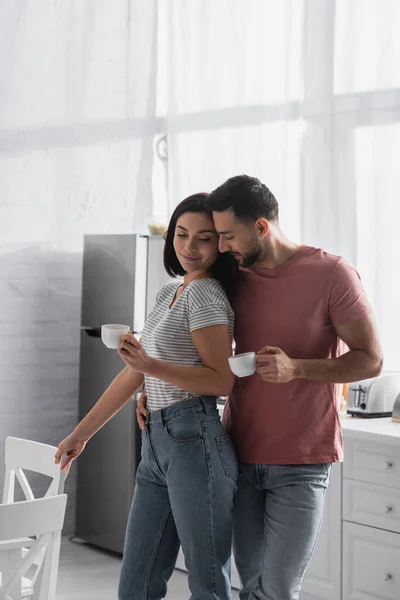 Feliz joven pareja abrazándose con tazas de café cerca de sillas en la cocina - foto de stock