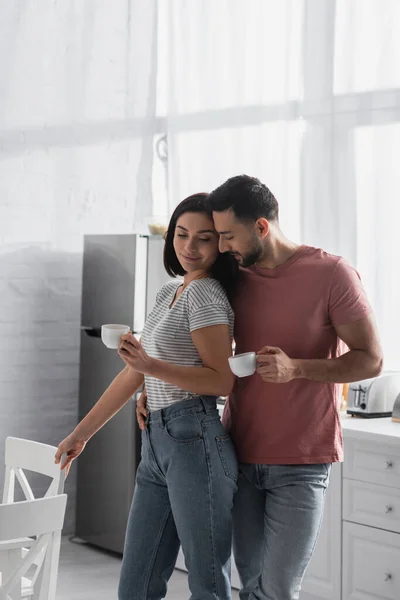 Heureux jeune couple étreignant avec tasses à café près de chaises dans la cuisine — Photo de stock