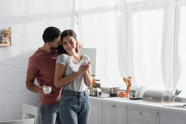 Feliz pareja joven abrazos y besos con tazas de café en la cocina - foto de stock