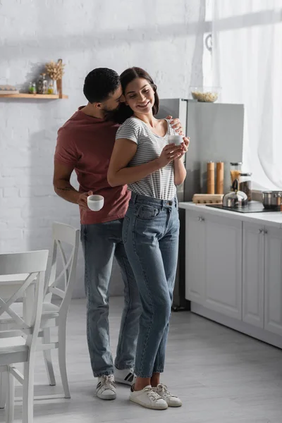 Sorrindo jovem casal abraçando e beijando com xícaras de café perto da mesa e cadeiras na cozinha — Fotografia de Stock