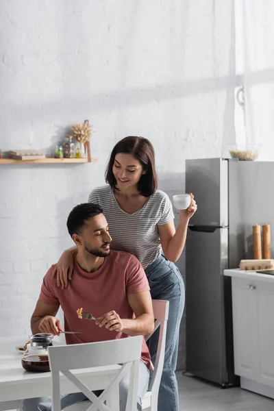Sorrindo jovem mulher abraçando namorado com pedaços de panquecas em garfo e segurando xícara com café na cozinha — Fotografia de Stock