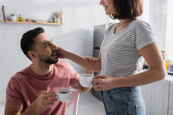 Young couple gently touching each other and holding cups with coffee in kitchen — Stock Photo