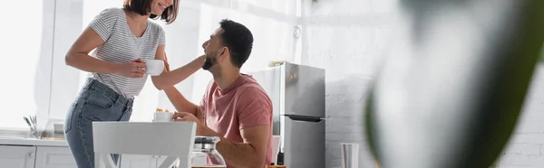 Young couple gently touching each other and holding cups with coffee in kitchen, banner — Stock Photo