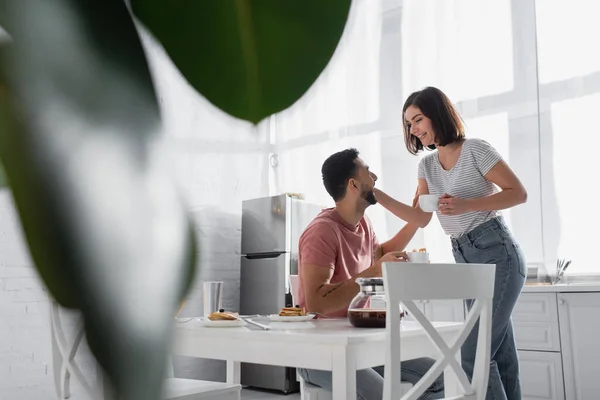 Pareja joven tocándose suavemente y sosteniendo tazas con café cerca de la mesa en la cocina - foto de stock