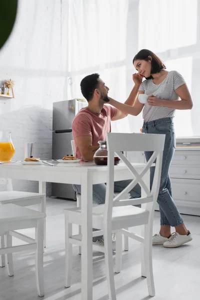 Молодая пара нежно прикасаясь друг к другу возле стола с завтраком и кофе на кухне — стоковое фото
