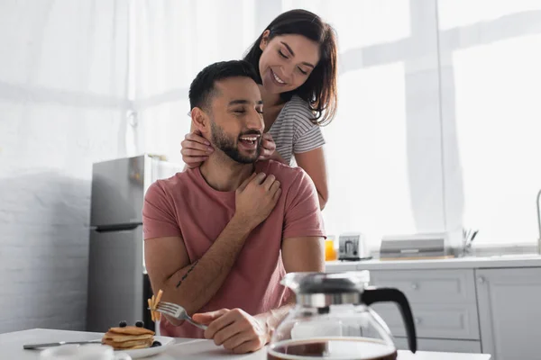 Smiling young woman gently touching boyfriend near table with breakfast and coffee in kitchen — Stock Photo