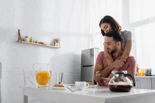 Lächelnde junge Frau umarmt Freund am Tisch mit Frühstück, Orangensaft und Kaffee in der Küche — Stockfoto