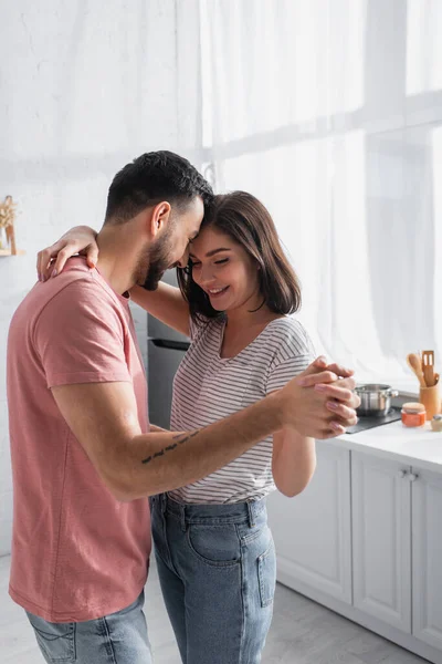 Улыбающаяся молодая пара в футболках и джинсах, танцующая на кухне — стоковое фото