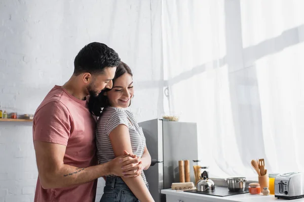 Sonriente joven pareja enamorada gentilmente abrazándose en la cocina - foto de stock