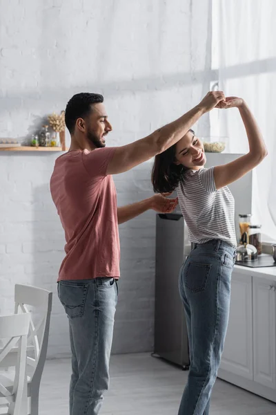 Positive young couple in casual clothes dancing with holding hands in modern kitchen — Stock Photo