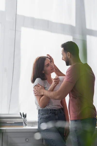 Smiling young man gently touching girlfriend with white coffee cup in modern kitchen — Stock Photo