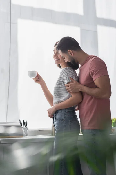 Усміхнений молодий чоловік ніжно цілує дівчину з чашкою білої кави на сучасній кухні — стокове фото