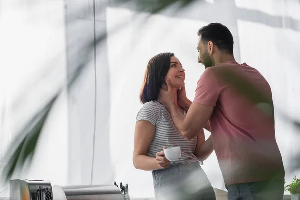 Giovane uomo sorridente toccando collo di fidanzata con tazza di caffè bianco in cucina moderna — Foto stock