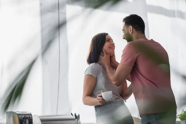 Smiling young man touching neck of girlfriend with white coffee cup in modern kitchen — Stock Photo