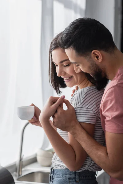 Mujer joven positiva con taza de café tocando las manos con el novio en la cocina moderna - foto de stock