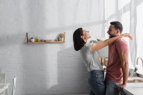 Sonriente joven pareja mirándose y abrazándose en la cocina - foto de stock
