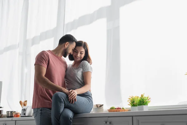 Улыбающийся молодой человек обнимает девушку с рукой на бедре на кухне — стоковое фото