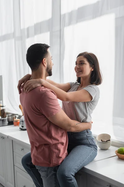 Sonriente joven pareja mirándose y abrazándose en la cocina - foto de stock