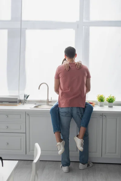 Back view of young man hugging girlfriend sitting on cupboard in kitchen — Stock Photo