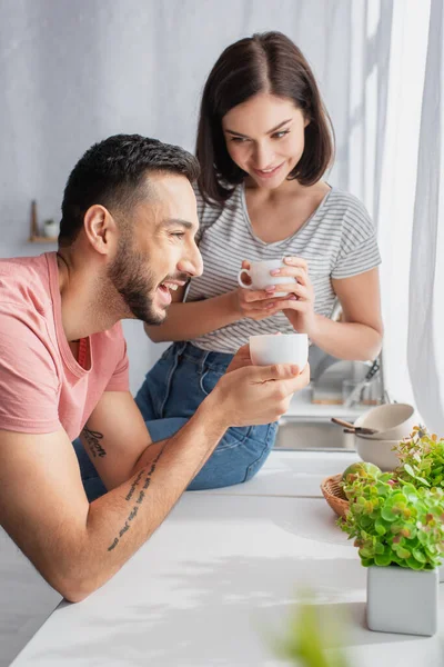Sonriente pareja joven sosteniendo tazas blancas con café cerca de la ventana en la cocina - foto de stock
