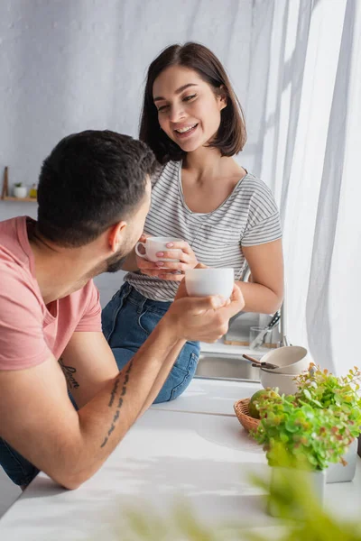 Jovem casal positivo segurando copos brancos com café perto da janela na cozinha — Fotografia de Stock