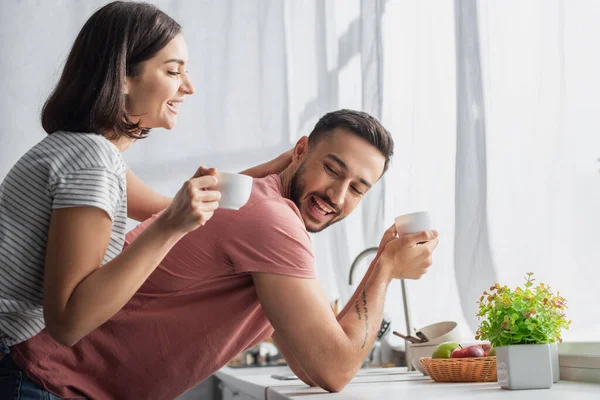 Smiling young woman holding white cup with coffee and touching boyfriend near window in kitchen — Stock Photo