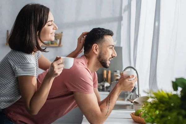 Positive young woman holding white cup with coffee and touching boyfriend near window in kitchen — Stock Photo