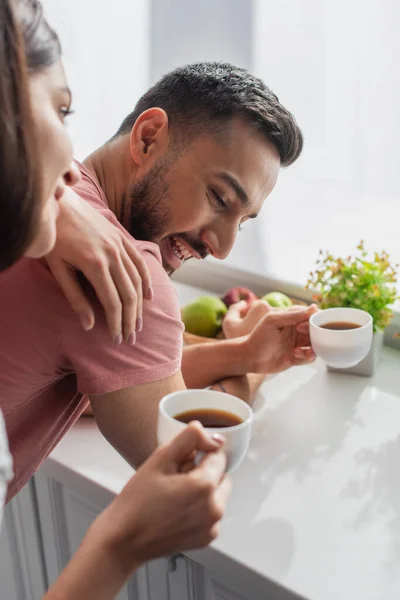 Sorrindo jovem segurando copo branco com café e namorado tocando perto da janela na cozinha — Fotografia de Stock
