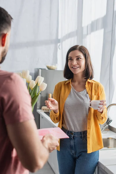 Hombre joven que presenta ramo de flores y tarjeta de felicitación a la novia en la cocina - foto de stock