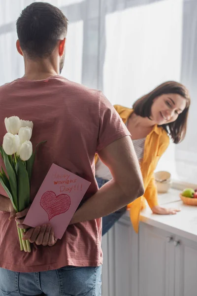 Vue arrière du jeune homme tenant bouquet de fleurs et carte de vœux près de petite amie floue dans la cuisine — Photo de stock